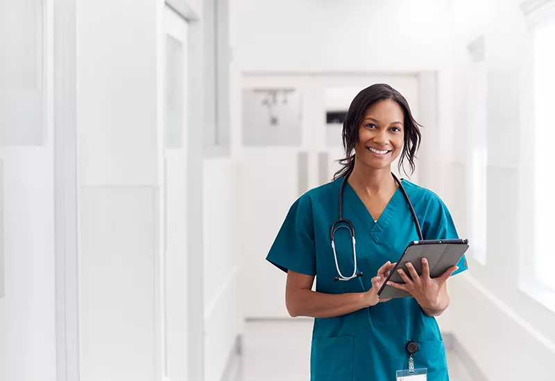 Portrait Of Smiling Female Doctor Wearing Scrubs In Hospital Corridor Holding Digital Tablet